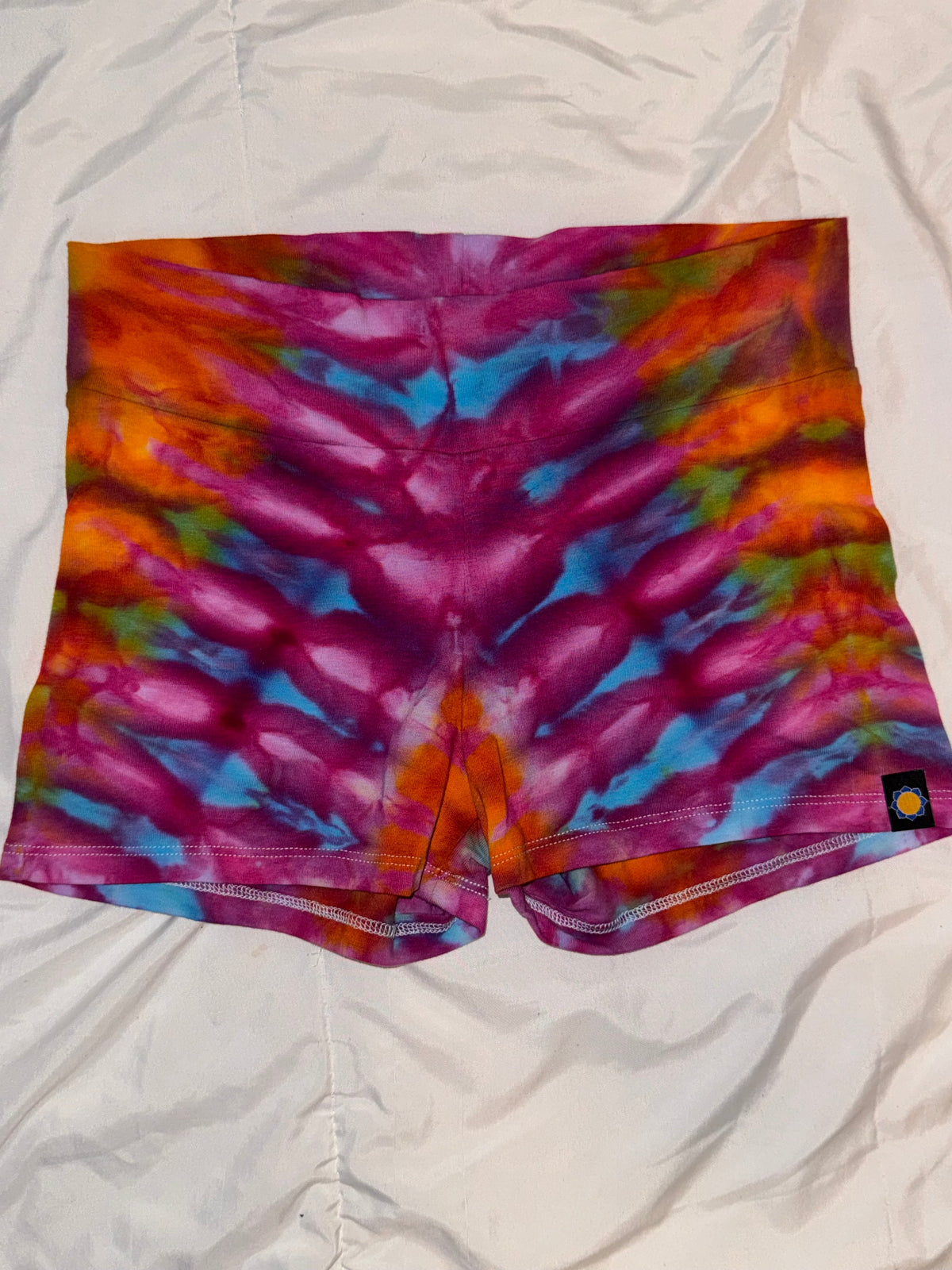 Violet Aura Booty Shorts in vibrant tie-dye — purple, orange, and blue swirl pattern, high-waist cut, festival-ready rave bottoms